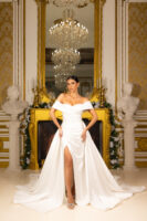 Elegant off-the-shoulder white wedding gown with draped bodice, pearl embellishments, high front slit, and flowing train, modeled in an opulent gold-accented interior.
