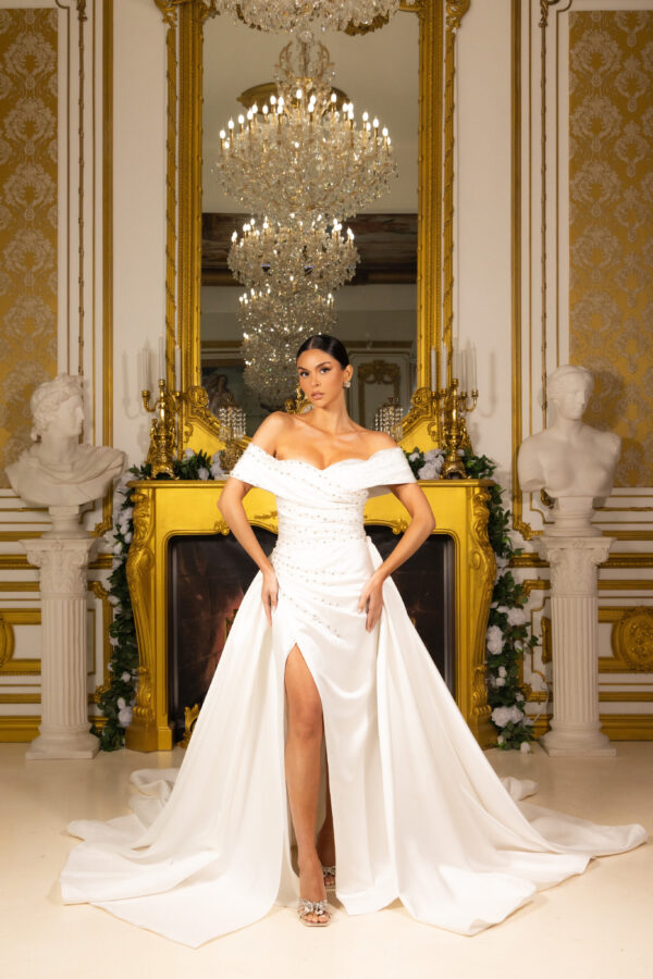 Elegant off-the-shoulder white wedding gown with draped bodice, pearl embellishments, high front slit, and flowing train, modeled in an opulent gold-accented interior.