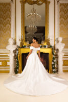 Elegant off-the-shoulder white ball gown wedding dress with a fitted bodice, full flowing train, and button details down the back, worn by a model standing in an opulent gold-accented room with a chandelier and classical busts.