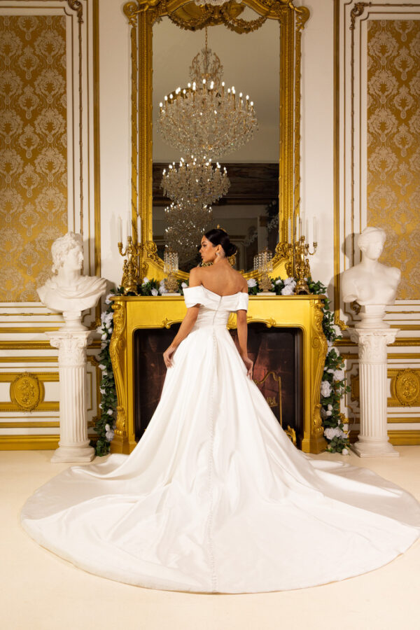 Elegant off-the-shoulder white ball gown wedding dress with a fitted bodice, full flowing train, and button details down the back, worn by a model standing in an opulent gold-accented room with a chandelier and classical busts.