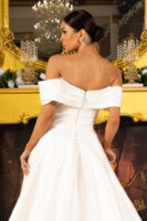 Back view of a model wearing an elegant off-the-shoulder white wedding gown with draped bodice, fitted waist, exposed zipper, and full A-line skirt featuring button details, set against a luxurious ornate fireplace and chandelier backdrop.
