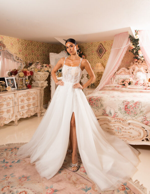 Model wearing an elegant white ballgown wedding dress with delicate lace bodice, thin straps, and a flowing tulle skirt featuring a high front slit, posed confidently in a luxurious floral-decorated bedroom.
