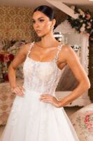 Model wearing an elegant white strapless wedding gown with delicate 3D floral appliqués, lace embroidery, and beadwork on the bodice, featuring thin straps adorned with leaf-like embellishments, posed confidently in a floral-decorated room.