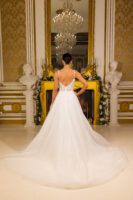 Rear view of a bride in an elegant white ball gown wedding dress with delicate lace appliqués on the sheer bodice, thin straps, and a voluminous tulle skirt with a long cathedral train, posed in an opulent room with gold accents and a chandelier.