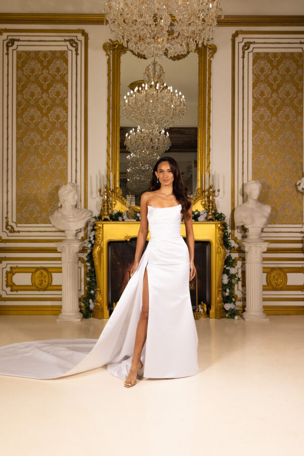 Elegant strapless white wedding gown with a fitted bodice, high side slit, and flowing train, modeled in an opulent room with gold accents and chandelier.