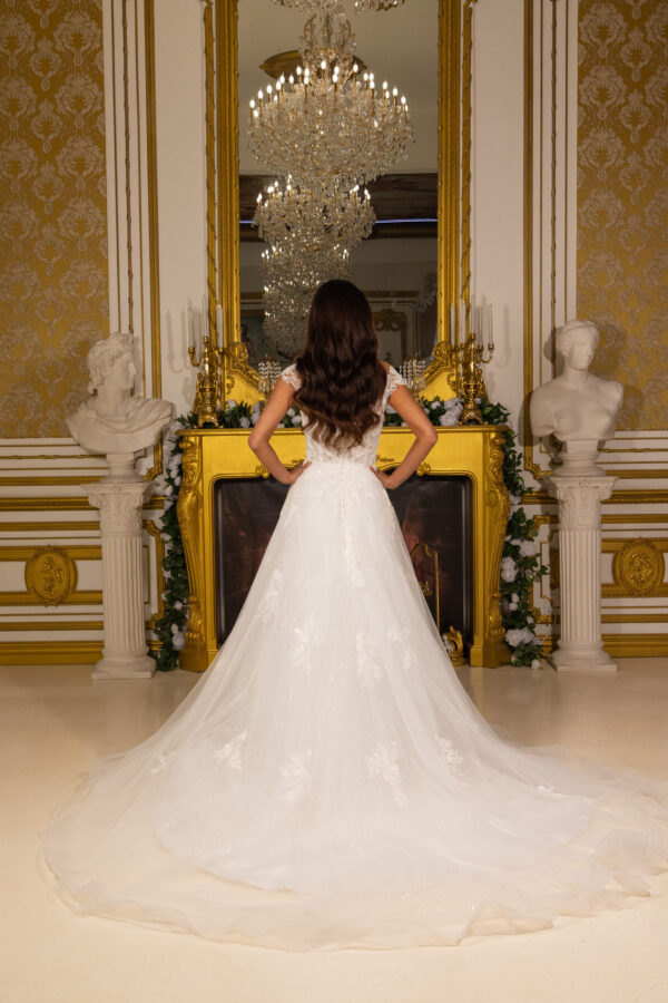 Rear view of a bride in an elegant off-the-shoulder white wedding gown with intricate lace appliqués, a glittering tulle skirt, and a long cathedral train, posing in a luxurious ornate room with gold accents and a chandelier.