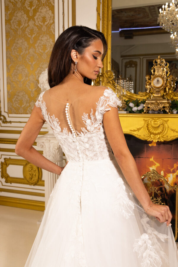 Model wearing an elegant white wedding gown with intricate floral lace appliqués on the cap sleeves and sheer illusion back, featuring a row of covered buttons down the spine, in a luxurious ornate interior.