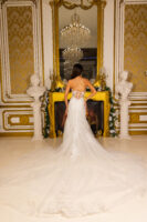 Back view of a bride in a strapless white wedding gown with intricate floral lace appliqués, sheer corset-style bodice with buttons, and a flowing cathedral-length train, posed in an opulent room with gold accents and a chandelier.