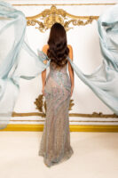 Rear view of a model wearing an elegant off-the-shoulder evening gown in shimmering pastel blue-green with intricate sequin and bead embellishments in swirling patterns, holding flowing sheer fabric drapes against an ornate gold-decorated backdrop.