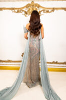 Model wearing an elegant, floor-length evening gown in shimmering sage green and nude tones with intricate sequin and bead embroidery in swirling patterns, featuring off-the-shoulder straps and long flowing chiffon cape sleeves, viewed from behind against an ornate backdrop.