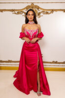 Elegant off-the-shoulder fuchsia satin evening gown with intricate silver crystal embroidery on the bodice, draped chains, puffed sleeves, high side slit, and matching embellished choker, modeled against an ornate gold-accented wall.