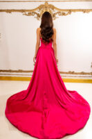 Model wearing a vibrant hot pink strapless evening gown with a fitted bodice featuring corset-style lacing at the back and a voluminous floor-length ball skirt with a dramatic train, viewed from behind against an ornate gold-accented wall.