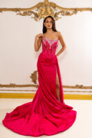 Model wearing a vibrant fuchsia strapless evening gown with intricate silver beadwork and crystal embellishments on the bodice, featuring draped chains and a high side slit, with a long flowing train.