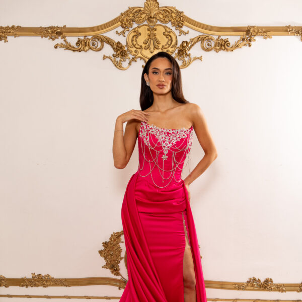 Model wearing a vibrant fuchsia strapless evening gown with intricate silver beadwork and crystal embellishments on the bodice, featuring draped chains and a high side slit, with a long flowing train.