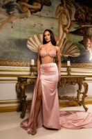 Model wearing a glamorous blush pink evening gown featuring a strapless sheer corset bodice with embellished shell-shaped cups and boning details, paired with a flowing satin skirt with a high side slit and train, posed in front of Botticelli's Birth of Venus painting.