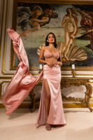 Model wearing a luxurious pink satin gown featuring a strapless embellished bustier top with pearl and bead detailing, paired with a high-slit flowing skirt and dramatic draped train, posed in front of Botticelli's Birth of Venus painting.