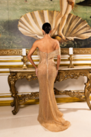 Model wearing a shimmering gold off-the-shoulder evening gown with corset-style lace-up back detailing, viewed from behind in an ornate setting with a classical painting backdrop.