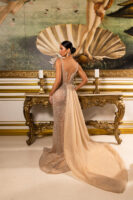 Model wearing an elegant champagne-colored evening gown with intricate sequin and bead embellishments, sheer overlay, corset-style lace-up back, flowing chiffon train, and bow shoulder detail, posed in profile against a classical Venus painting backdrop.