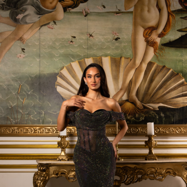 Model wearing a sparkling black off-the-shoulder gown with sheer mesh bodice and floor-length sequined skirt, posed in front of Botticelli's Birth of Venus painting in an ornate gold-framed setting.