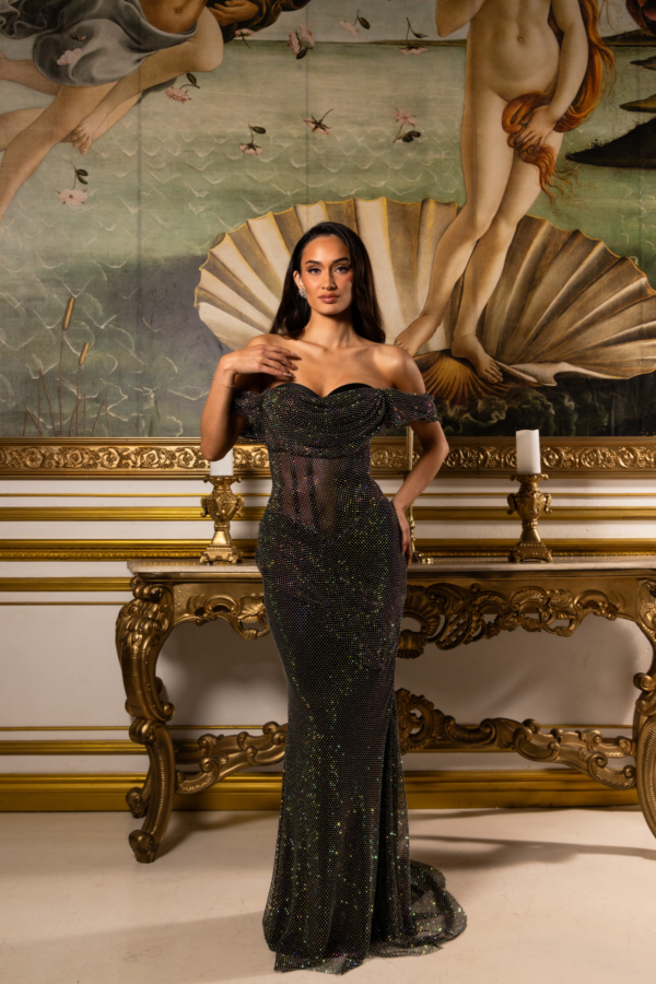Model wearing a sparkling black off-the-shoulder gown with sheer mesh bodice and floor-length sequined skirt, posed in front of Botticelli's Birth of Venus painting in an ornate gold-framed setting.