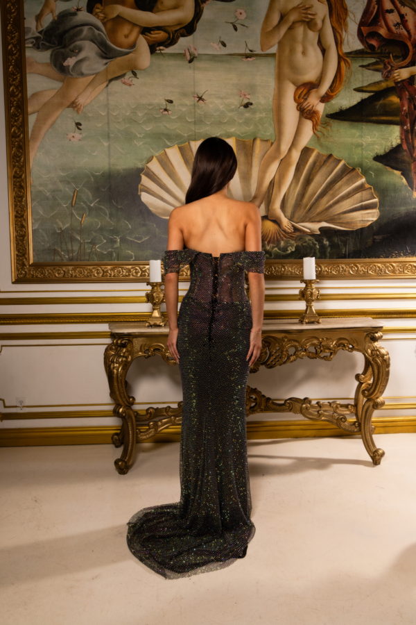 Rear view of a model wearing a sparkling black off-the-shoulder evening gown with iridescent sequins, corset-style lace-up back, fitted mermaid silhouette, and flowing train, standing before Botticelli's *The Birth of Venus* painting.