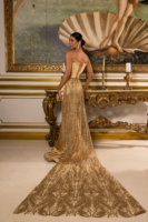 Elegant strapless gold evening gown with a fitted satin corset bodice and flowing sheer overlay skirt featuring intricate beadwork, sequins, and embroidery, complete with a dramatic long train. Model shown from behind in an opulent setting.