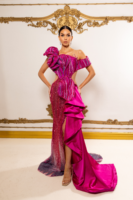 Model wearing a glamorous off-the-shoulder magenta evening gown with intricate sequin embellishments in vertical stripes, a large dramatic ruffle on one shoulder, high side slit, and flowing asymmetrical train.