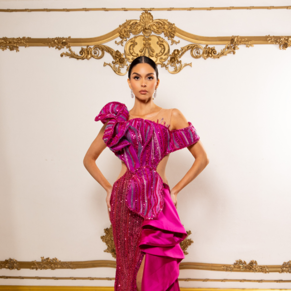 Model wearing a glamorous off-the-shoulder magenta evening gown with intricate sequin embellishments in vertical stripes, a large dramatic ruffle on one shoulder, high side slit, and flowing asymmetrical train.