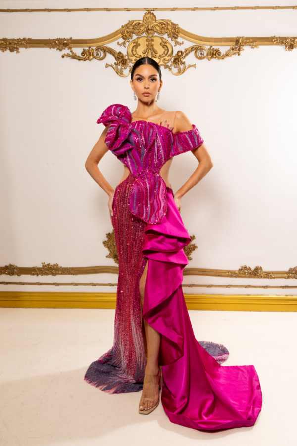 Model wearing a glamorous off-the-shoulder magenta evening gown with intricate sequin embellishments in vertical stripes, a large dramatic ruffle on one shoulder, high side slit, and flowing asymmetrical train.