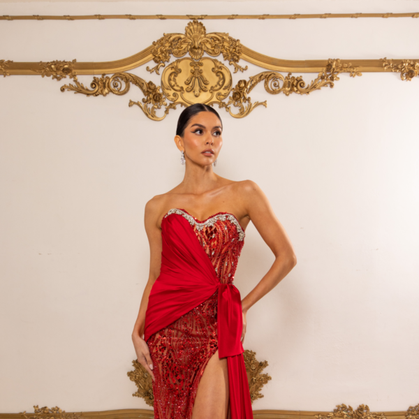Elegant red strapless evening gown with intricate sequin and bead embellishments on a sheer overlay, featuring an asymmetrical draped sash, high side slit, and flowing train.