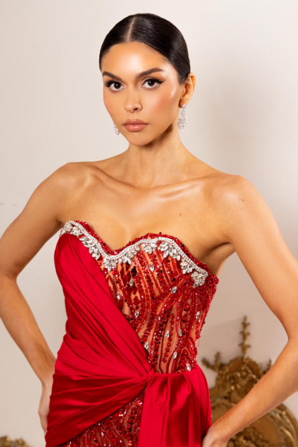 Model wearing a strapless red evening gown with intricate crystal and sequin embellishments on the bodice, draped asymmetric fabric, and elegant dangling earrings.