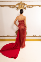 Model wearing a strapless red sequined evening gown with intricate embellishments and a long flowing train, viewed from behind against an ornate gold-decorated wall.