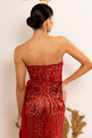 Rear view of a model wearing a strapless red evening gown with intricate sequin and bead embroidery in swirling patterns, sheer mesh panels, fitted mermaid silhouette, and a draped fabric accent at the waist.