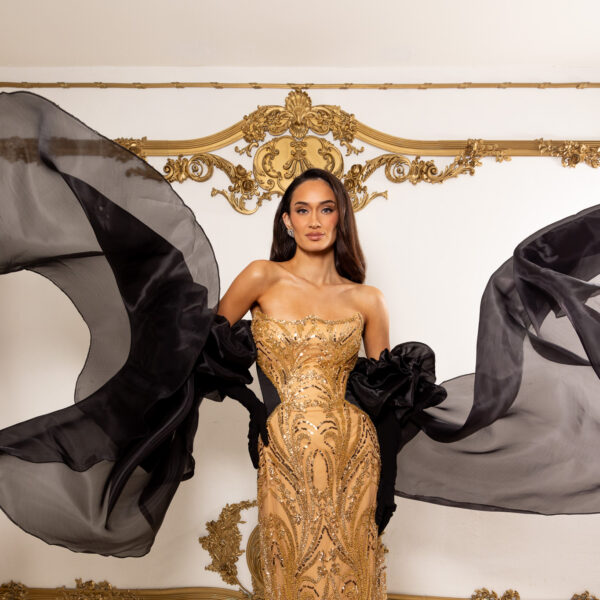 Model wearing an elegant strapless gold gown with intricate sequin and bead embroidery in swirling patterns, featuring dramatic flowing black chiffon cape sleeves, posed against an ornate gold-accented wall.
