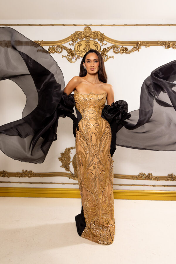 Model wearing an elegant strapless gold gown with intricate sequin and bead embroidery in swirling patterns, featuring dramatic flowing black chiffon cape sleeves, posed against an ornate gold-accented wall.