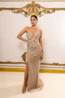 Model wearing a luxurious nude illusion evening gown with intricate silver beadwork and floral appliqués, featuring a sheer bodice, high slit, and floor-length train against an ornate gold-accented backdrop.