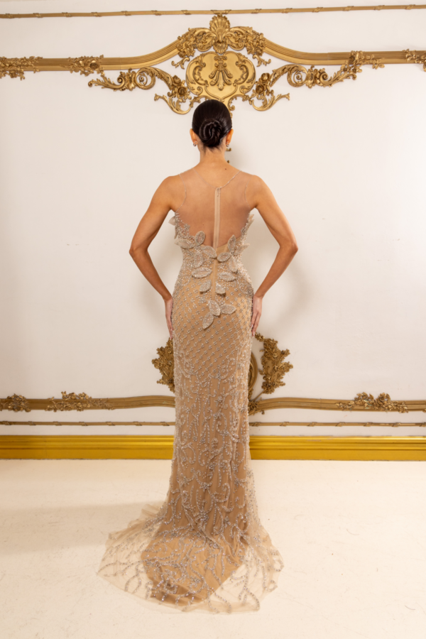 Back view of a model wearing an elegant, floor-length champagne-colored evening gown with intricate silver beadwork and floral embroidery, sheer illusion back panels, and a subtle train, set against an ornate gold-accented wall.