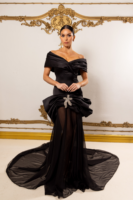 Model wearing an elegant black off-the-shoulder evening gown with draped bodice, peplum detail, crystal floral brooch at the waist, sheer overlay, and flowing train.