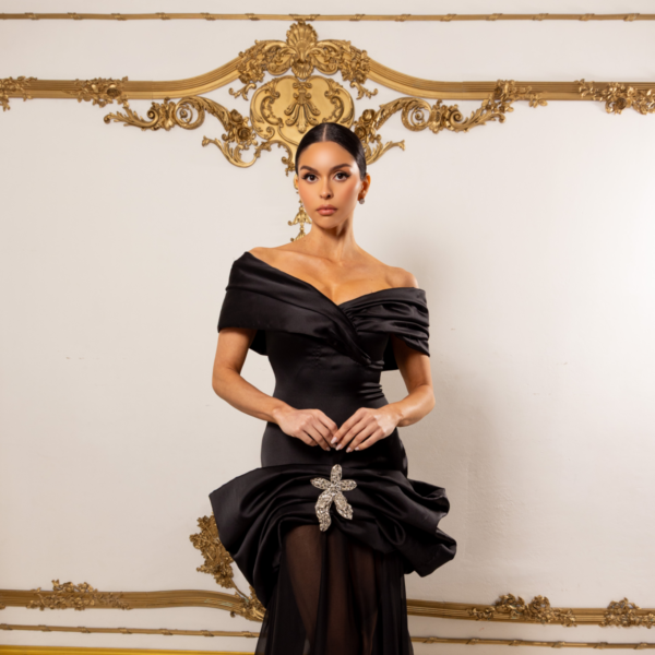 Model wearing an elegant black off-the-shoulder evening gown with draped bodice, peplum detail, crystal floral brooch at the waist, sheer overlay, and flowing train.