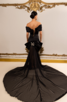 Rear view of a model wearing an elegant black off-the-shoulder evening gown with corset-style lace-up back, dramatic peplum detail, sparkling butterfly brooch at the waist, and flowing sheer train.