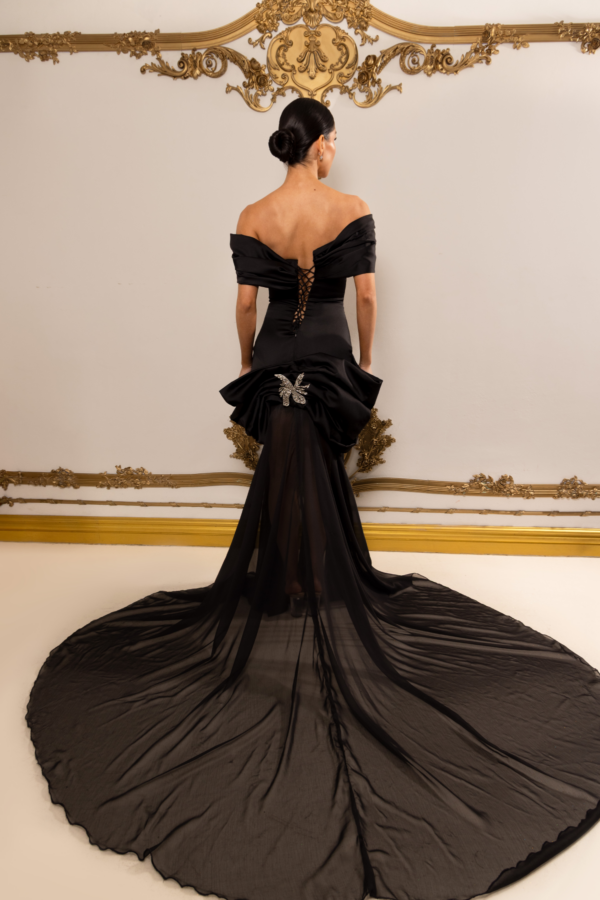 Rear view of a model wearing an elegant black off-the-shoulder evening gown with corset-style lace-up back, dramatic peplum detail, sparkling butterfly brooch at the waist, and flowing sheer train.
