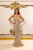 Model wearing a strapless, floor-length nude evening gown with intricate silver beadwork, embroidery, and crystal embellishments in swirling patterns, posed confidently with hands on hips against an ornate gold-trimmed backdrop.