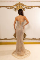 Rear view of a model wearing a strapless champagne-colored mermaid evening gown with intricate silver beadwork and embroidery throughout, featuring a corset-style lace-up back and a flowing train.