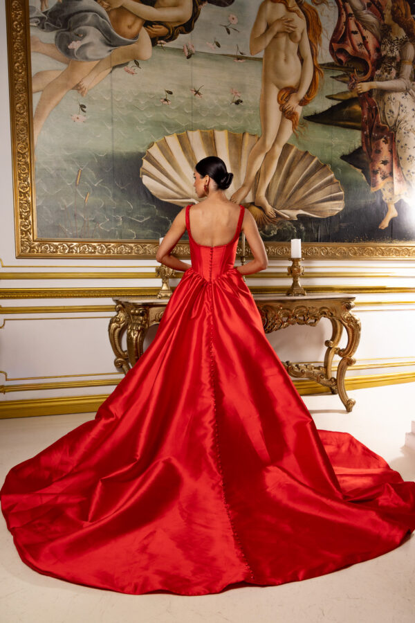 Elegant red satin ball gown with corset-style back featuring button detailing down the center, wide shoulder straps, and a voluminous floor-length train with chapel sweep, worn by a model facing away in front of Botticelli's Birth of Venus painting.