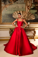Model wearing a luxurious red satin ball gown with a fitted corset bodice, wide straps, and a voluminous full-length skirt with train, posed in an opulent interior before a framed classical painting.