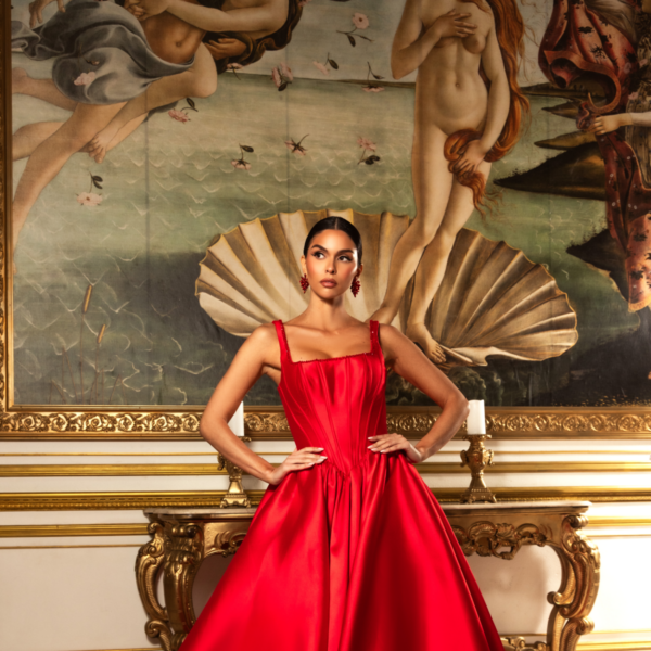 Model wearing an elegant, floor-length red satin ball gown with a fitted corset bodice, thick straps, and a voluminous skirt with train, posed confidently in front of a classical painting inspired by Botticelli's Birth of Venus.