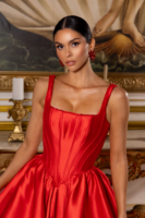 Model wearing a vibrant red satin ball gown with a fitted corset bodice featuring boning details, beaded square neckline and straps, paired with matching red floral earrings, posed in an opulent interior.
