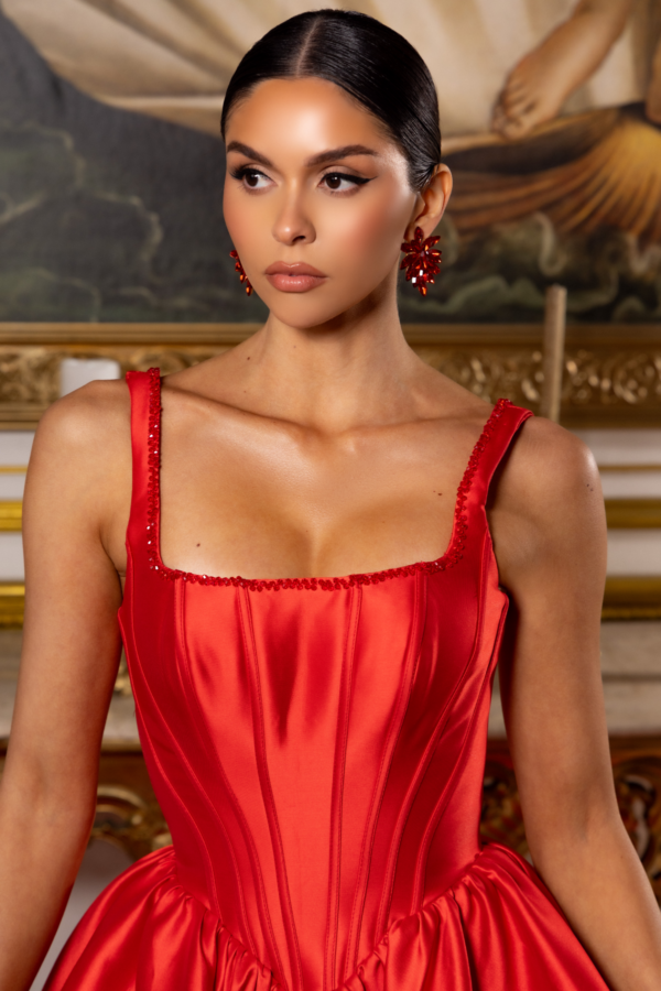 Elegant red satin corset gown with structured boning, square neckline adorned with red beading, thick straps, and a voluminous gathered skirt, modeled against a classical painted backdrop.