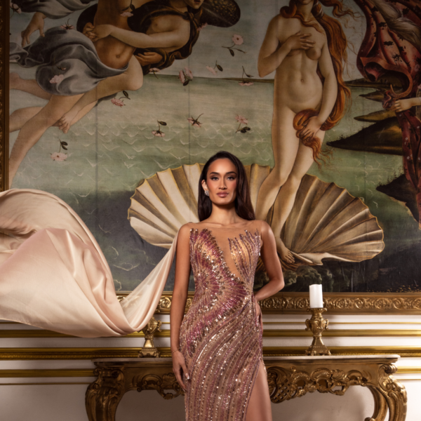 Model wearing a glamorous floor-length evening gown with intricate rose gold and pink sequin embellishments in swirling patterns, featuring a deep plunging neckline, high side slit, and flowing cape-like train, posed in front of a classical Renaissance-style painting.