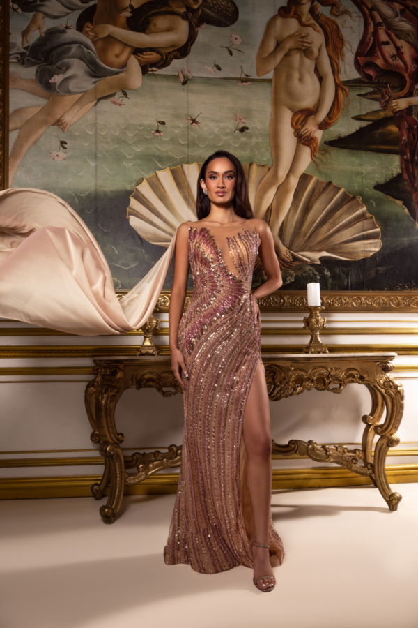 Model wearing a glamorous floor-length evening gown with intricate rose gold and pink sequin embellishments in swirling patterns, featuring a deep plunging neckline, high side slit, and flowing cape-like train, posed in front of a classical Renaissance-style painting.
